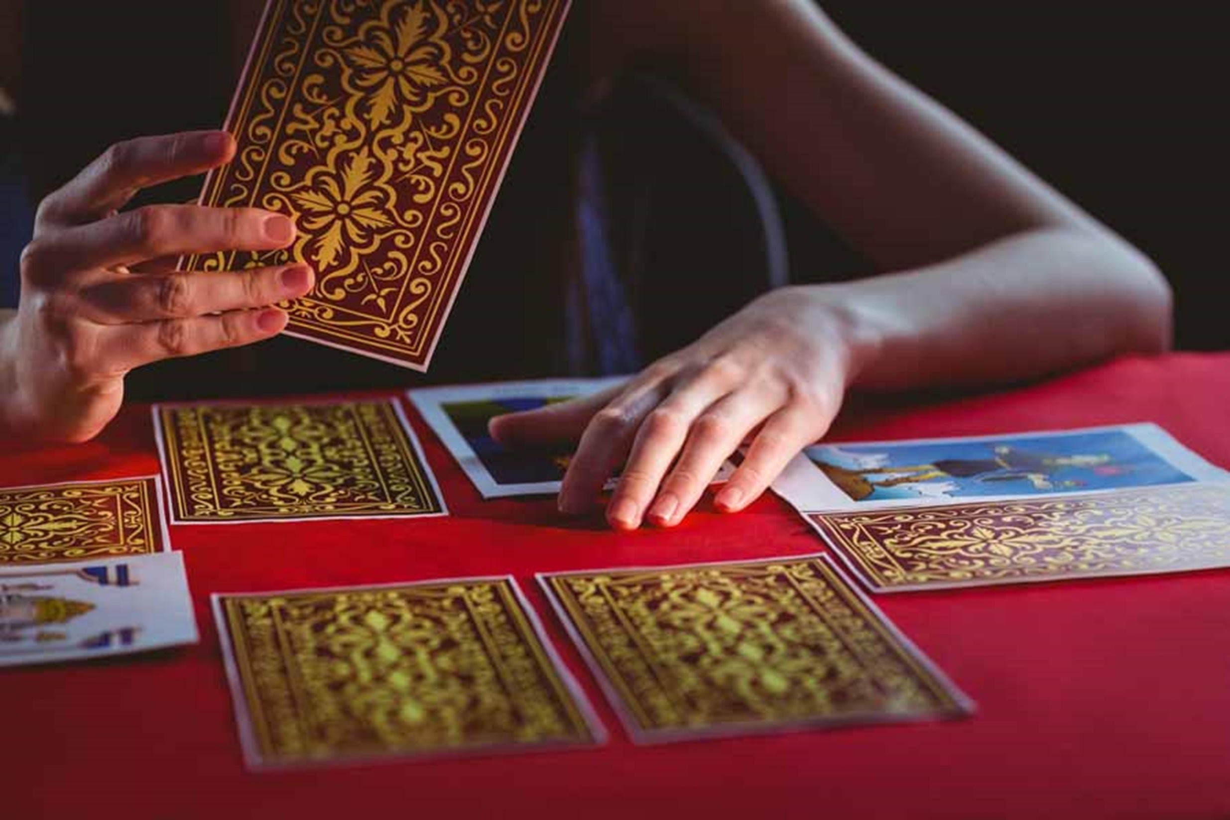 Torso y manos de una mujer joven. Está leyendo las cartas de tarot que están puestas sobre una mesa con mantel rojo. Hay varias cartas boca abajo y tiene una en su mano. 
